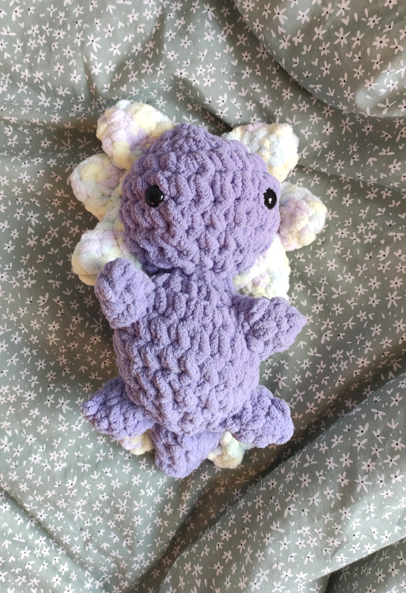 Ręcznie robiona, fioletowa, pluszowa ośmiornica z włóczki, leżąca na zielonym tle w białe kwiatki.