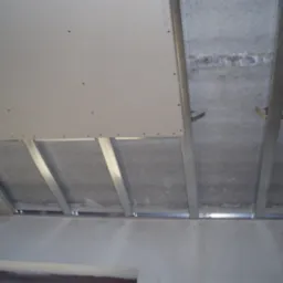 Sufit podwieszany w trakcie montażu, widoczne profile stalowe, płyta gipsowo-kartonowa częściowo zamontowana, surowy betonowy strop powyżej, ściany z widocznymi oznaczeniami pomiarowymi.