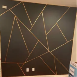 Ciemna ściana z geometrycznym wzorem z taśmy w kolorze złotym, widoczne gniazdka elektryczne i fragment podłogi z paneli.