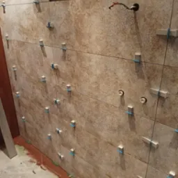 Ściana w trakcie układania płytek ceramicznych w odcieniach beżu i brązu, z widocznymi krzyżykami dystansowymi i sznurkiem wyznaczającym poziom, z wystającymi kablami elektrycznymi.