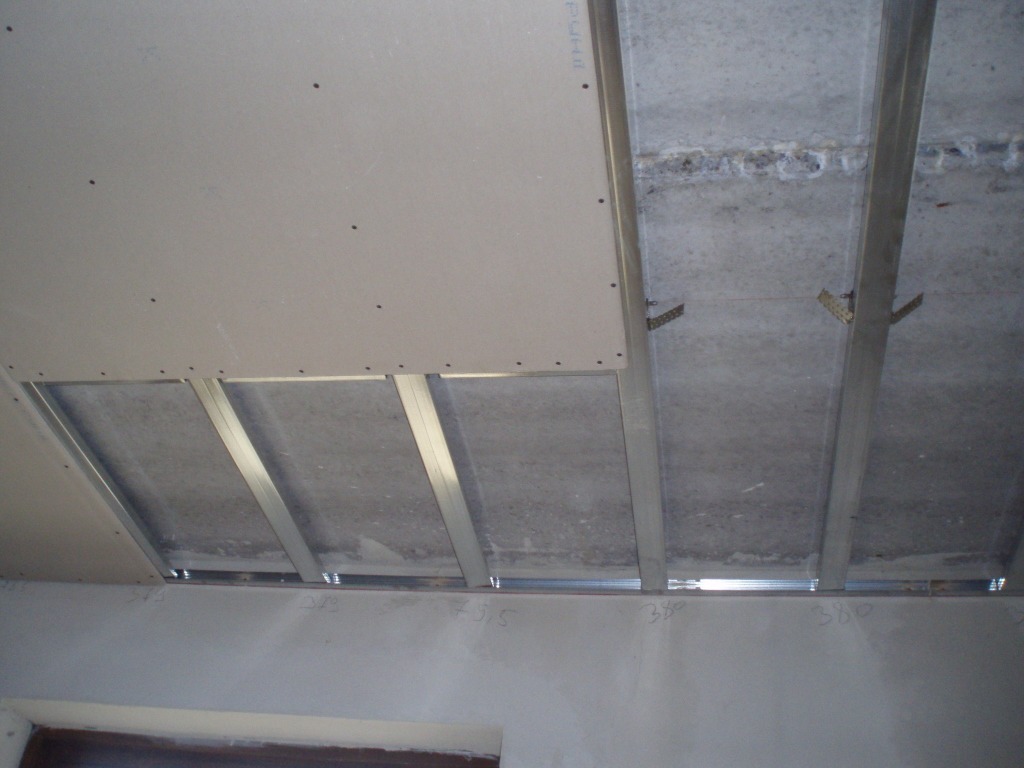 Sufit podwieszany w trakcie montażu, widoczne profile stalowe, płyta gipsowo-kartonowa częściowo zamontowana, surowy betonowy strop powyżej, ściany z widocznymi oznaczeniami pomiarowymi.