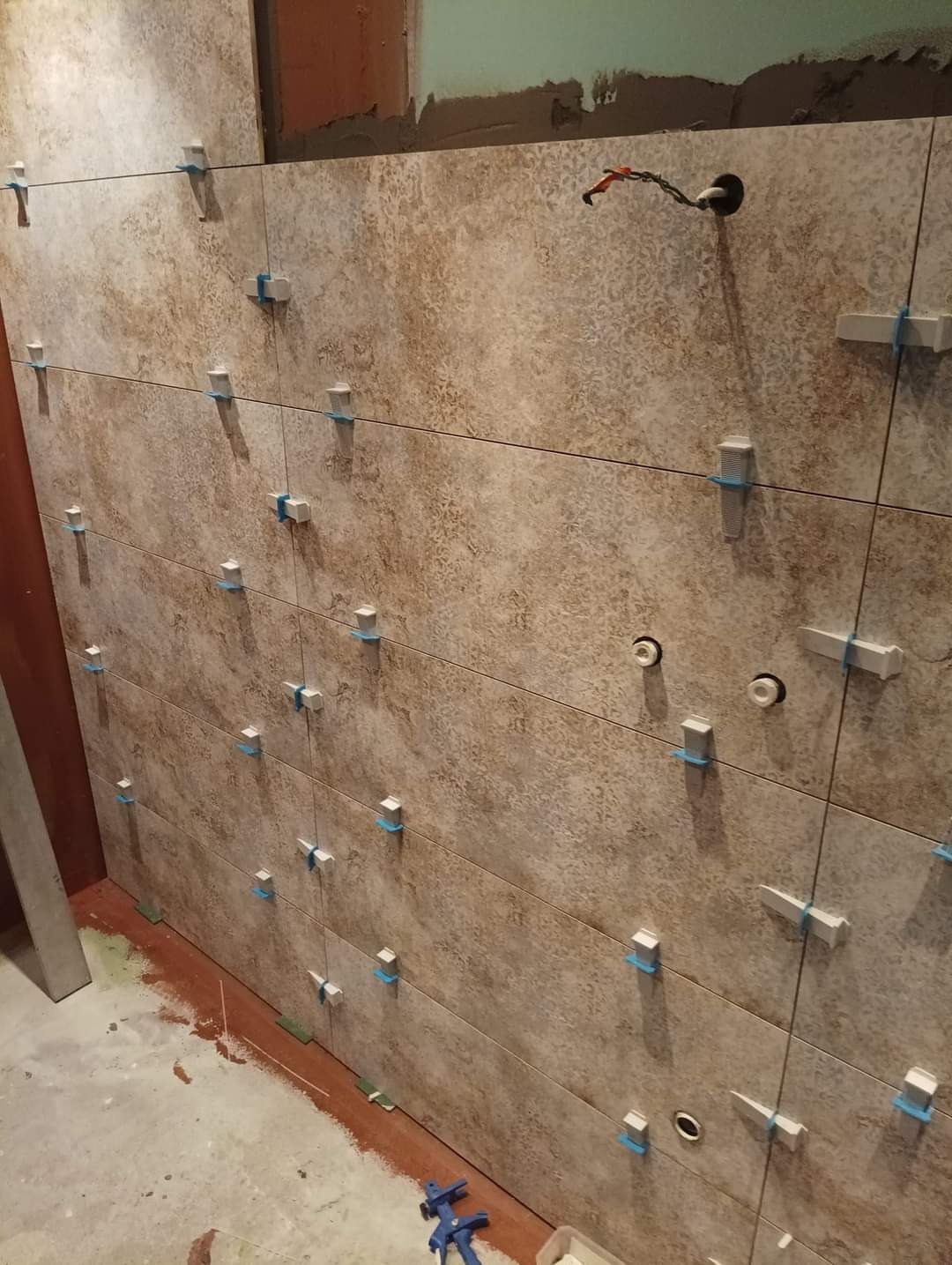 Ściana w trakcie układania płytek ceramicznych w odcieniach beżu i brązu, z widocznymi krzyżykami dystansowymi i sznurkiem wyznaczającym poziom, z wystającymi kablami elektrycznymi.