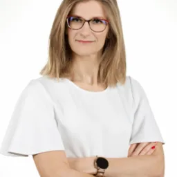 Portret kobiety w okularach z założonymi rękami, ubrana w białą bluzkę i spódnicę w kratę, z zegarkiem na ręce.