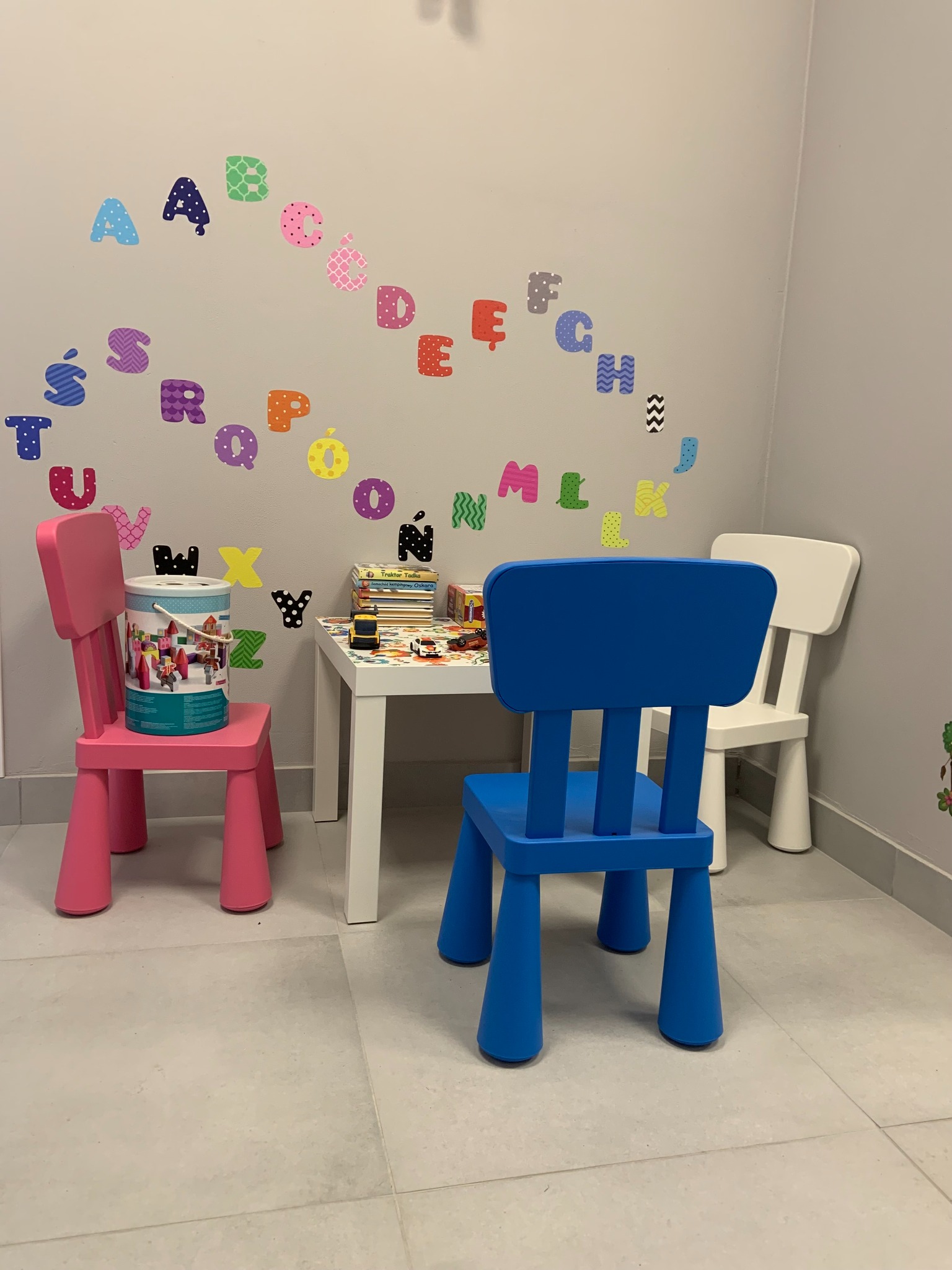 Pokój zabaw z kolorowymi krzesełkami dla dzieci, stolikiem z zabawkami i naklejkami z alfabetem na ścianie.