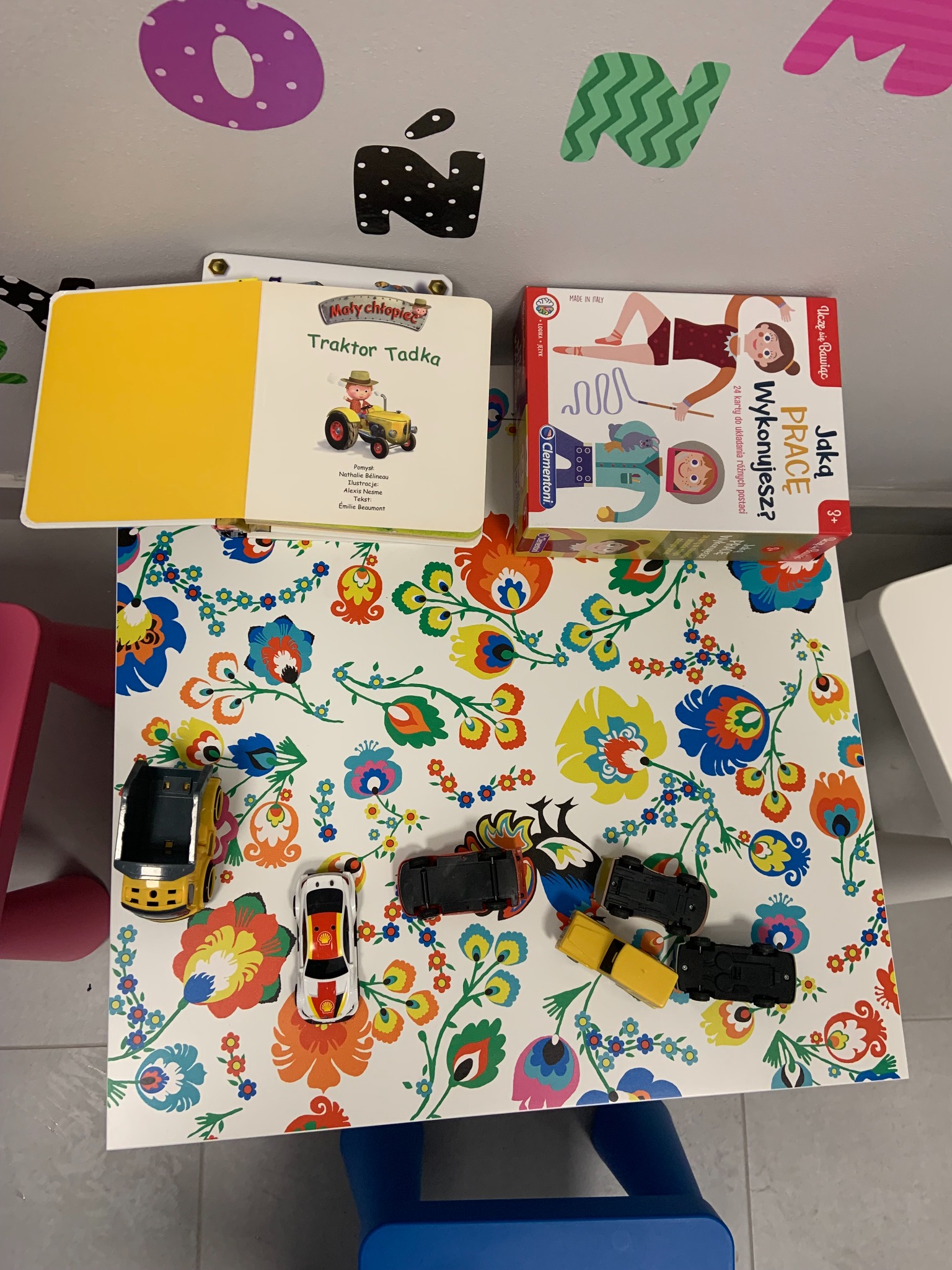 Książka dla dzieci 'Traktor Tadka' i gra edukacyjna 'Jaką pracę wykonujesz?' leżą na stoliku z kwiecistym wzorem, obok nich ustawione są zabawki - modele samochodów.