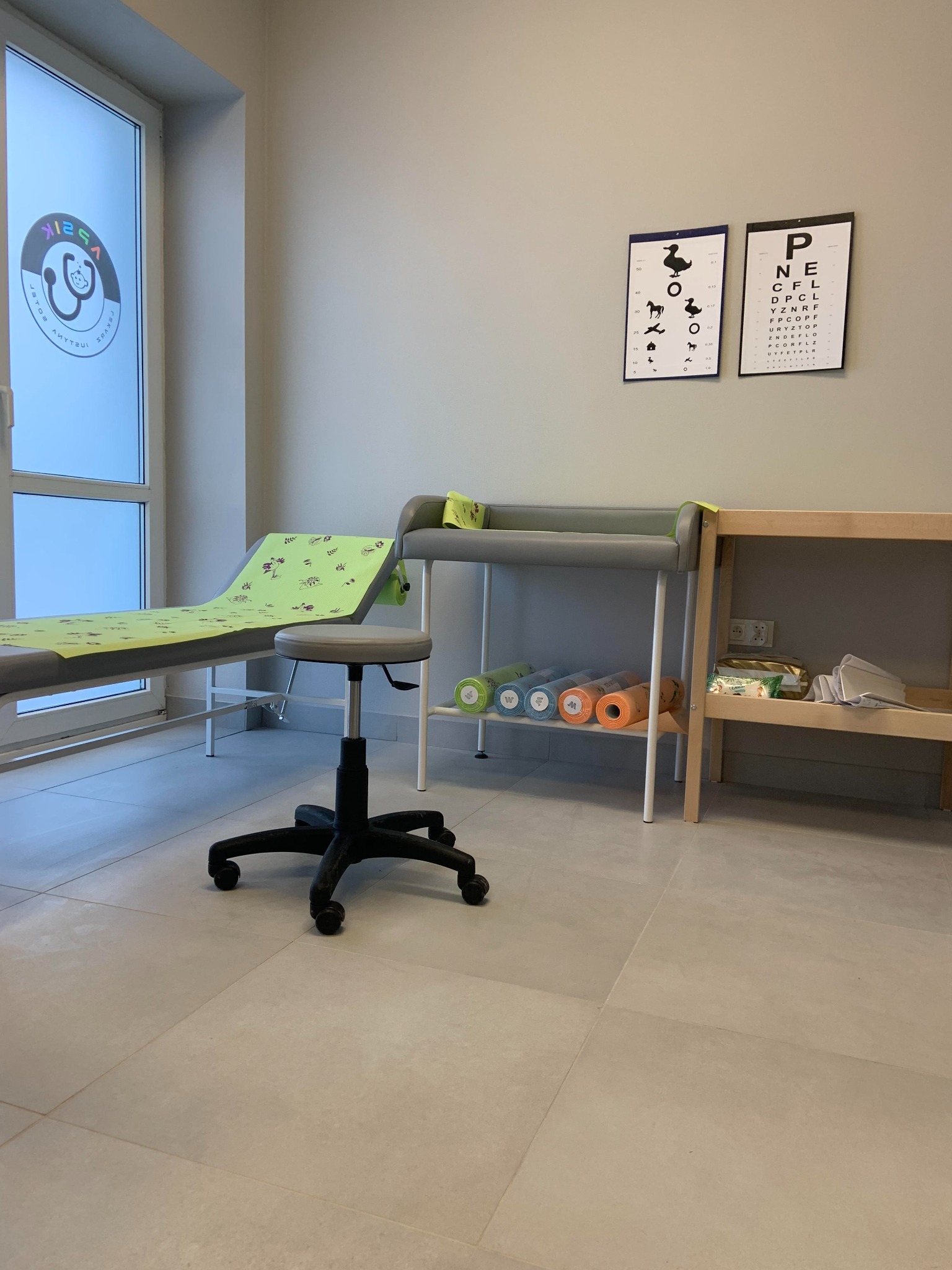 Gabinet lekarski w Lublinie: kozetka z zielonym podkładem w dziecięce wzory, tablica do badania wzroku z obrazkami zwierząt, przewijak z szarym materacem i stołek na kółkach.