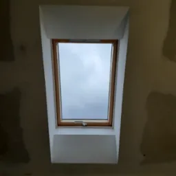 Zamontowane okno dachowe z drewnianą ramą i białą obudową w surowym, niepomalowanym wnętrzu poddasza.