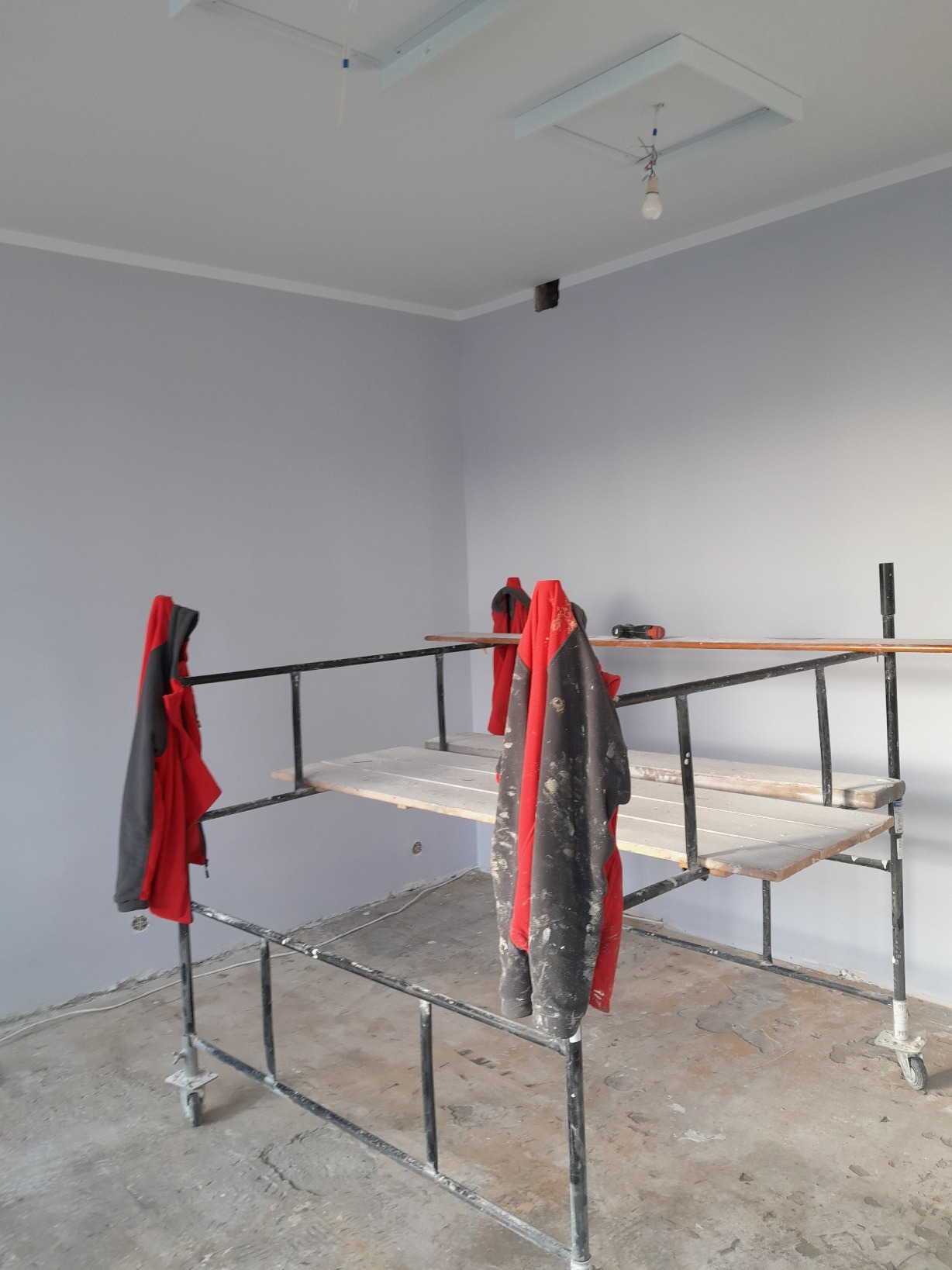 Pokój w trakcie malowania na szaro; widoczne rusztowanie z roboczymi ubraniami, sufit z otworem na oświetlenie i wystającymi kablami, oraz narzędzia na rusztowaniu.