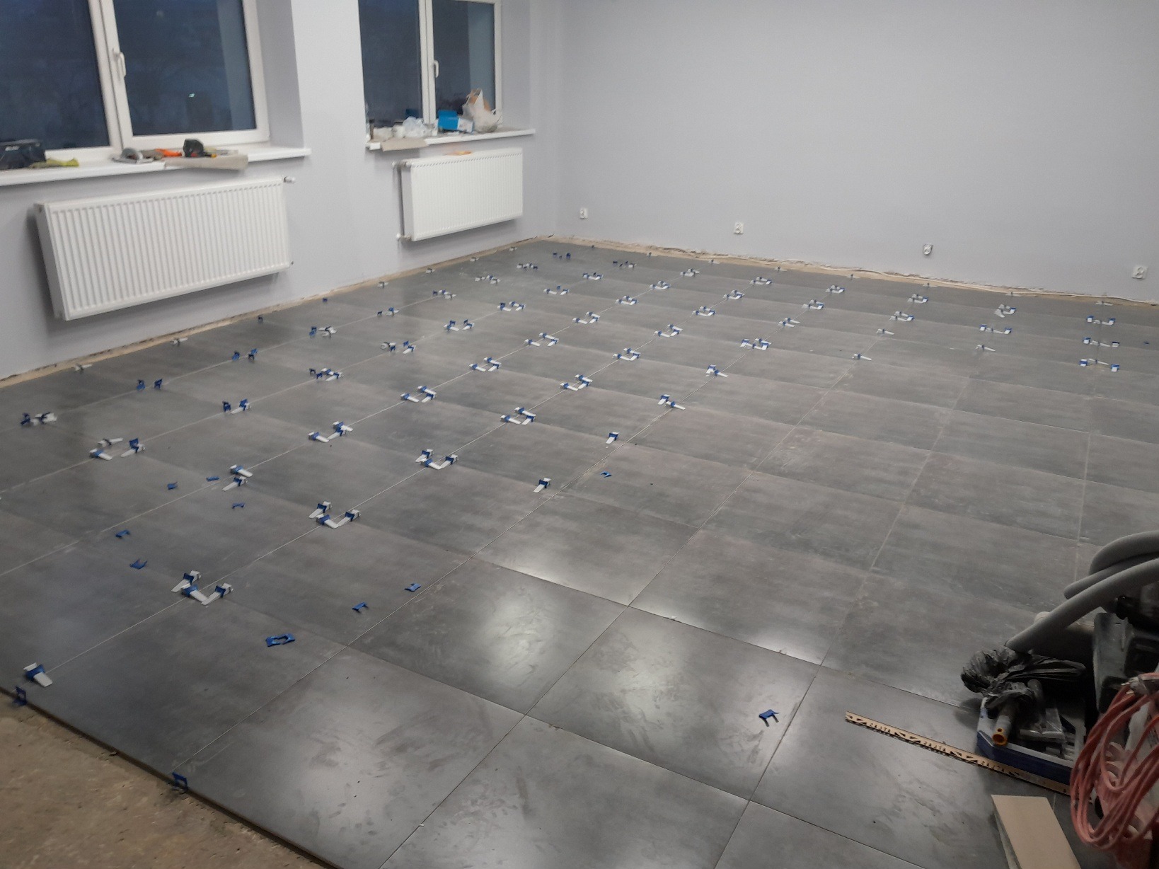 Szare płytki podłogowe w trakcie układania, z widocznymi niebieskimi krzyżykami dystansowymi, grzejniki na ścianie i odkurzacz przemysłowy w rogu pomieszczenia.