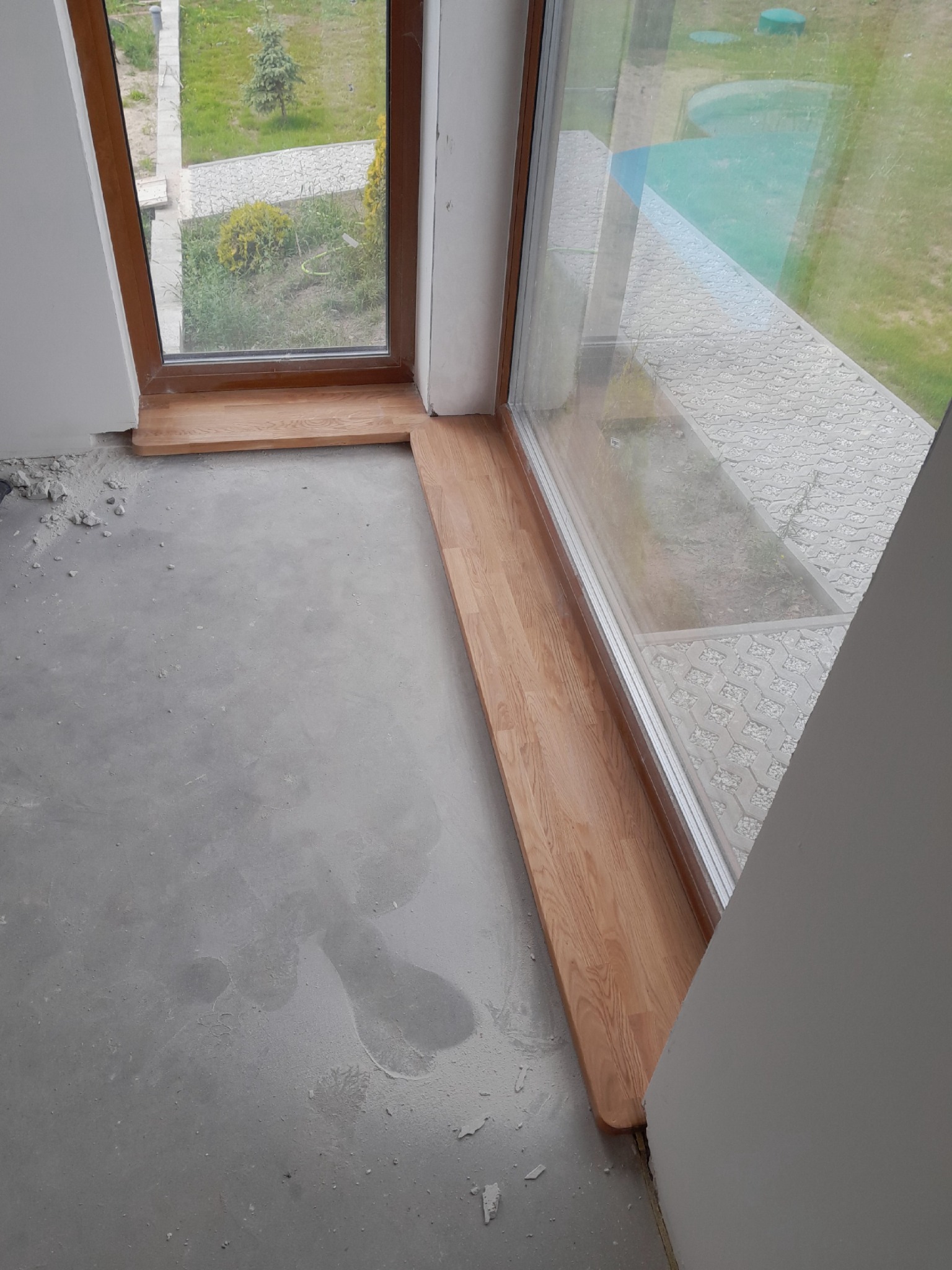 Montaż drewnianego parapetu wewnętrznego przy oknie z widokiem na trawnik i chodnik z ażurowych płyt. Surowy beton na podłodze, ślady po remoncie.