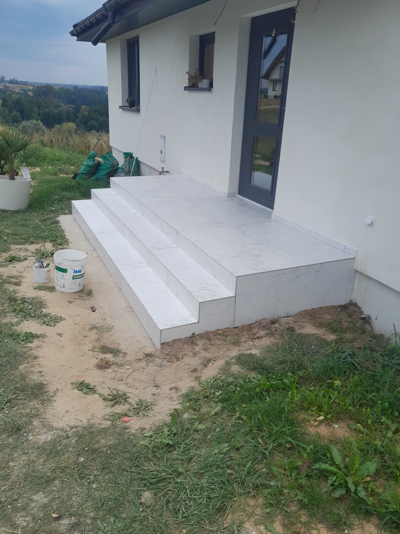 Wyłożone białymi płytkami schody wejściowe do domu z szarymi drzwiami, widoczne wiadro z klejem i narzędzia, w tle zieleń i budynki.