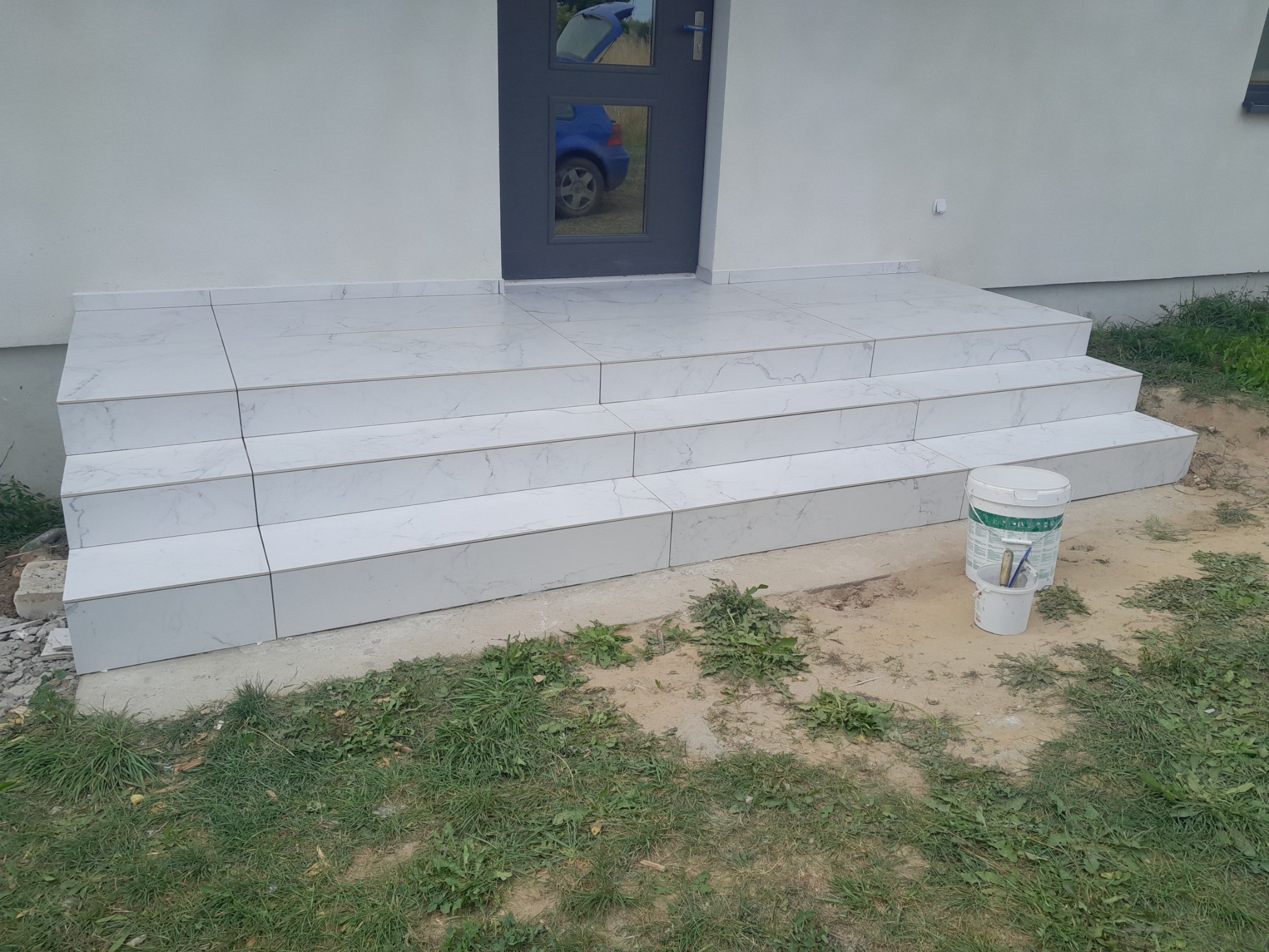 Wykończone płytkami schody wejściowe do domu, widoczny odbity w szybie samochód, obok wiadro z narzędziami.