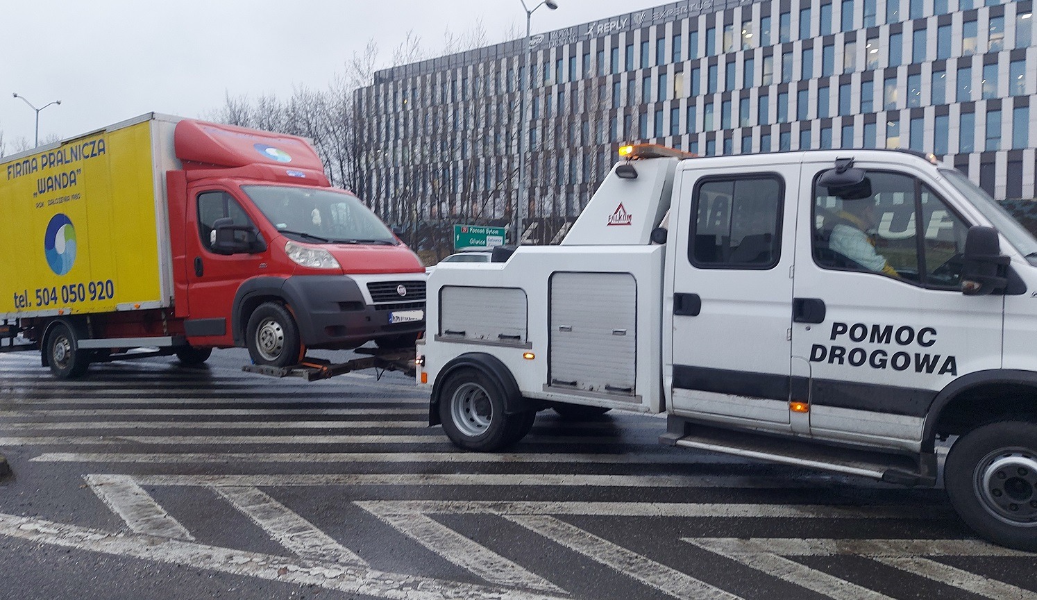 Biała ciężarówka pomocy drogowej marki Iveco z napisem 'POMOC DROGOWA' holuje na platformie czerwono-żółtego dostawczaka 'FIRMA PRALNICZA WANDA' na tle miejskiej zabudowy i mokrej jezdni.