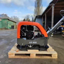 Nowa zagęszczarka gruntu LG 400 na drewnianej palecie, z pomarańczową ramą ochronną i silnikiem Honda GX, sfotografowana na zewnątrz w pochmurny dzień.