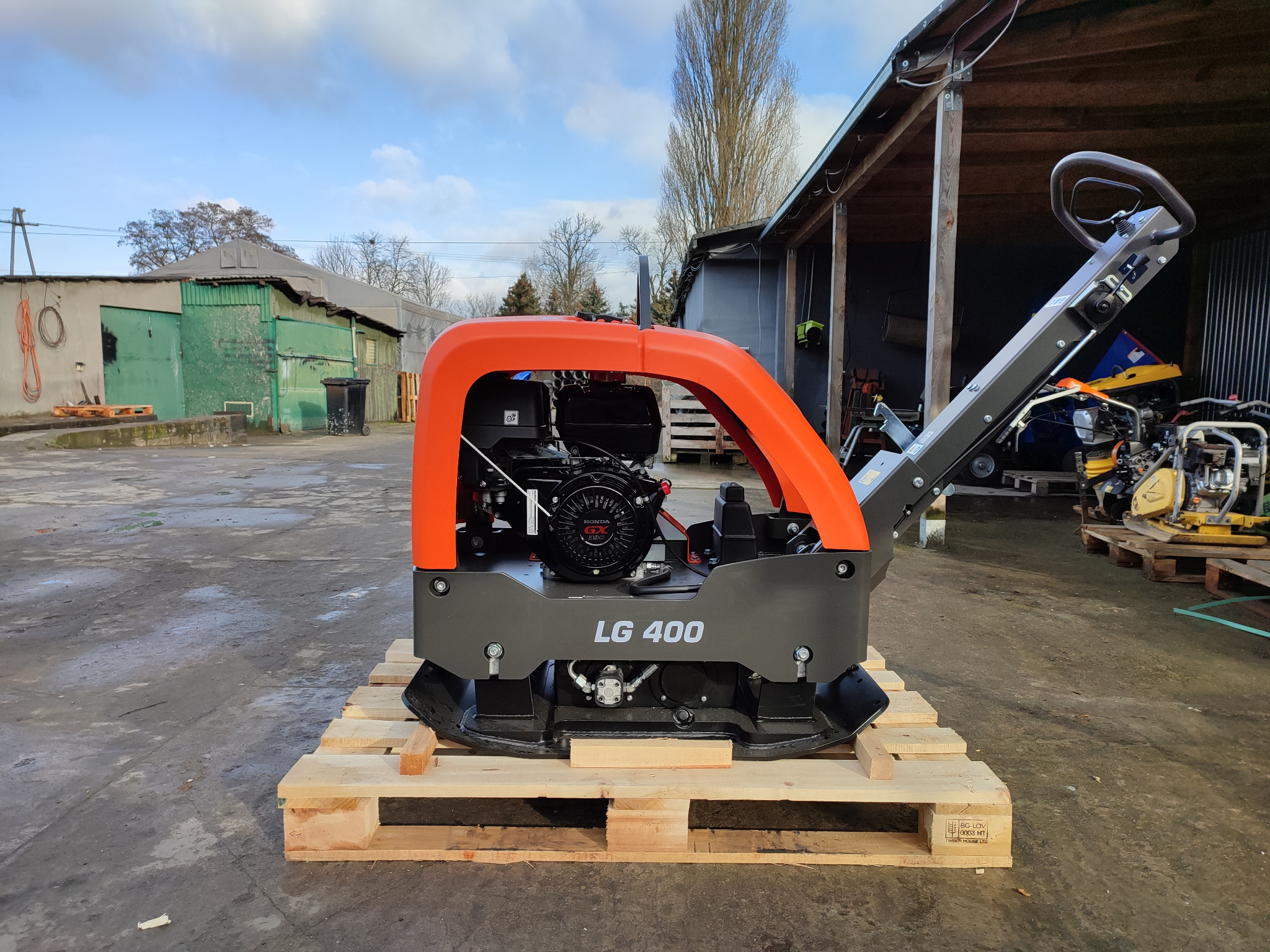 Nowa zagęszczarka gruntu LG 400 na drewnianej palecie, z pomarańczową ramą ochronną i silnikiem Honda GX, sfotografowana na zewnątrz w pochmurny dzień.