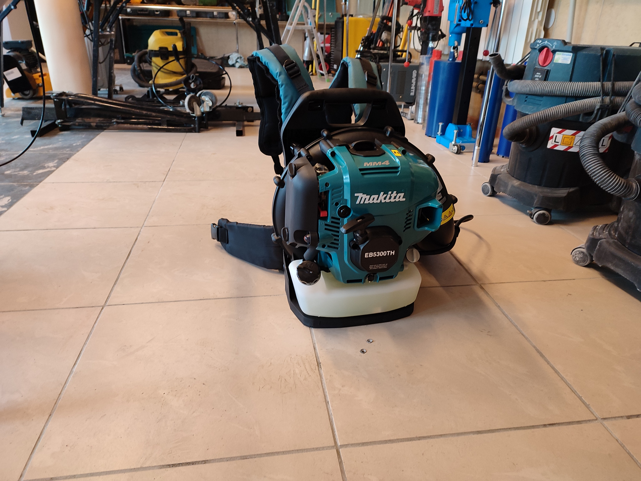 Plecakowy dmuchawa spalinowa Makita EBS5300TH w kolorze turkusowo-czarnym, stojąca na kafelkach w pomieszczeniu z widocznymi innymi maszynami i narzędziami budowlanymi w tle.