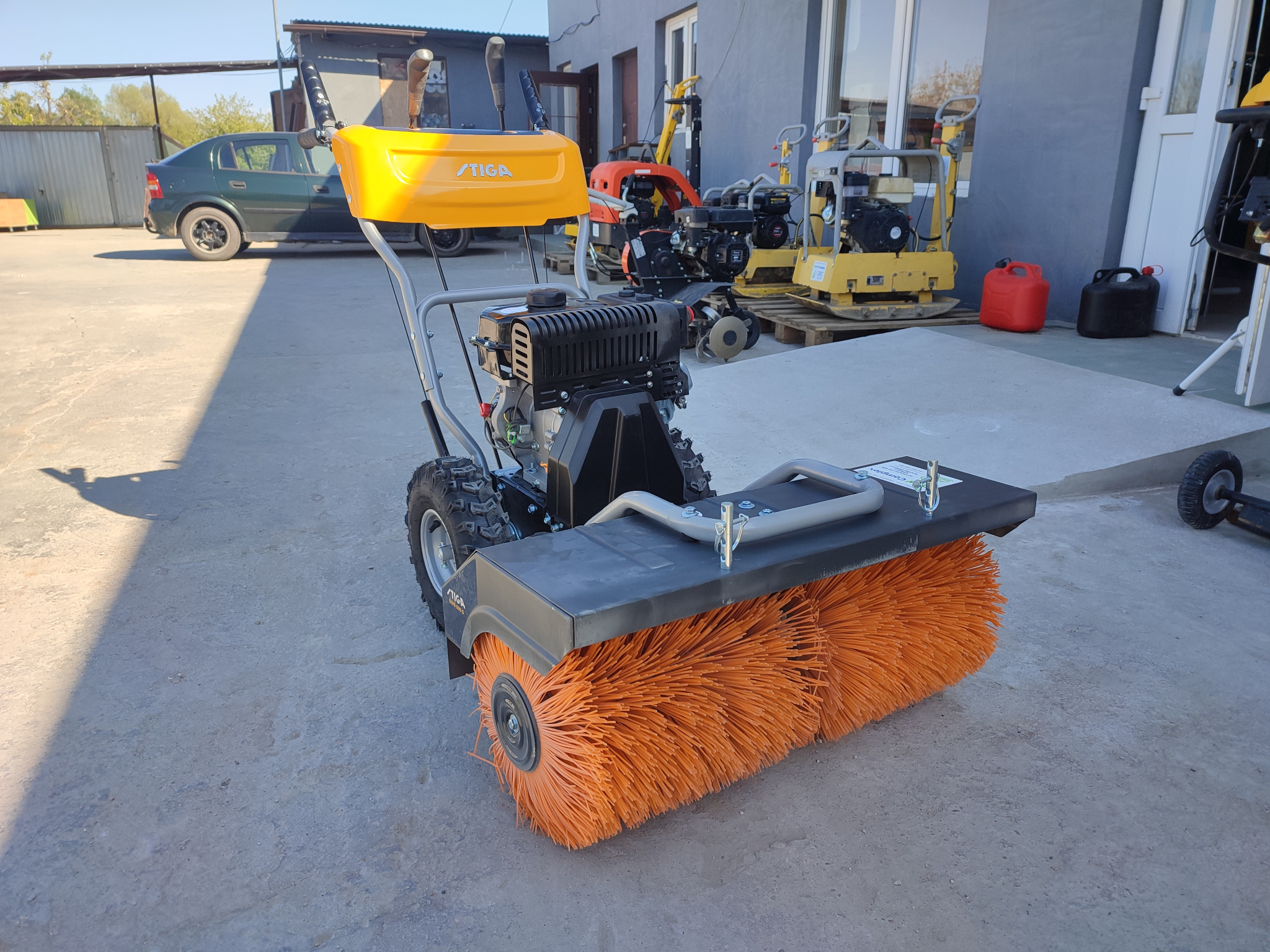 Żółto-szara zamiatarka spalinowa marki STIGA z pomarańczową szczotką walcową na betonowym podłożu, w tle inne maszyny budowlane i zielony samochód osobowy.