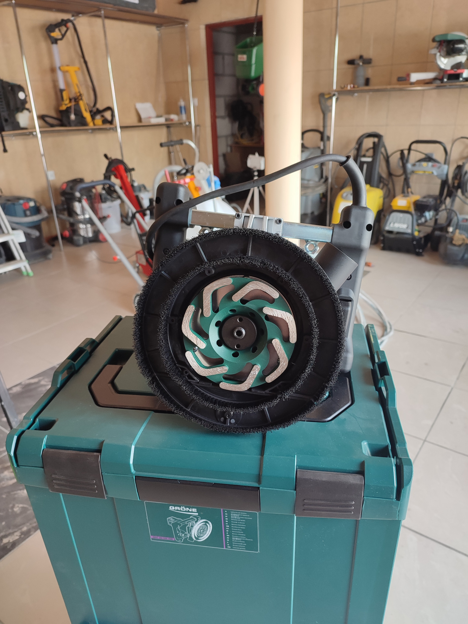 Szlifierka do betonu z tarczą szlifierską i szczotką, umieszczona na zielonym pojemniku transportowym marki Grone, w tle widoczne inne maszyny budowlane.