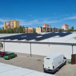 Rząd paneli słonecznych na dachu białego budynku z parkingiem, widok z góry w słoneczny dzień; w tle bloki mieszkalne.