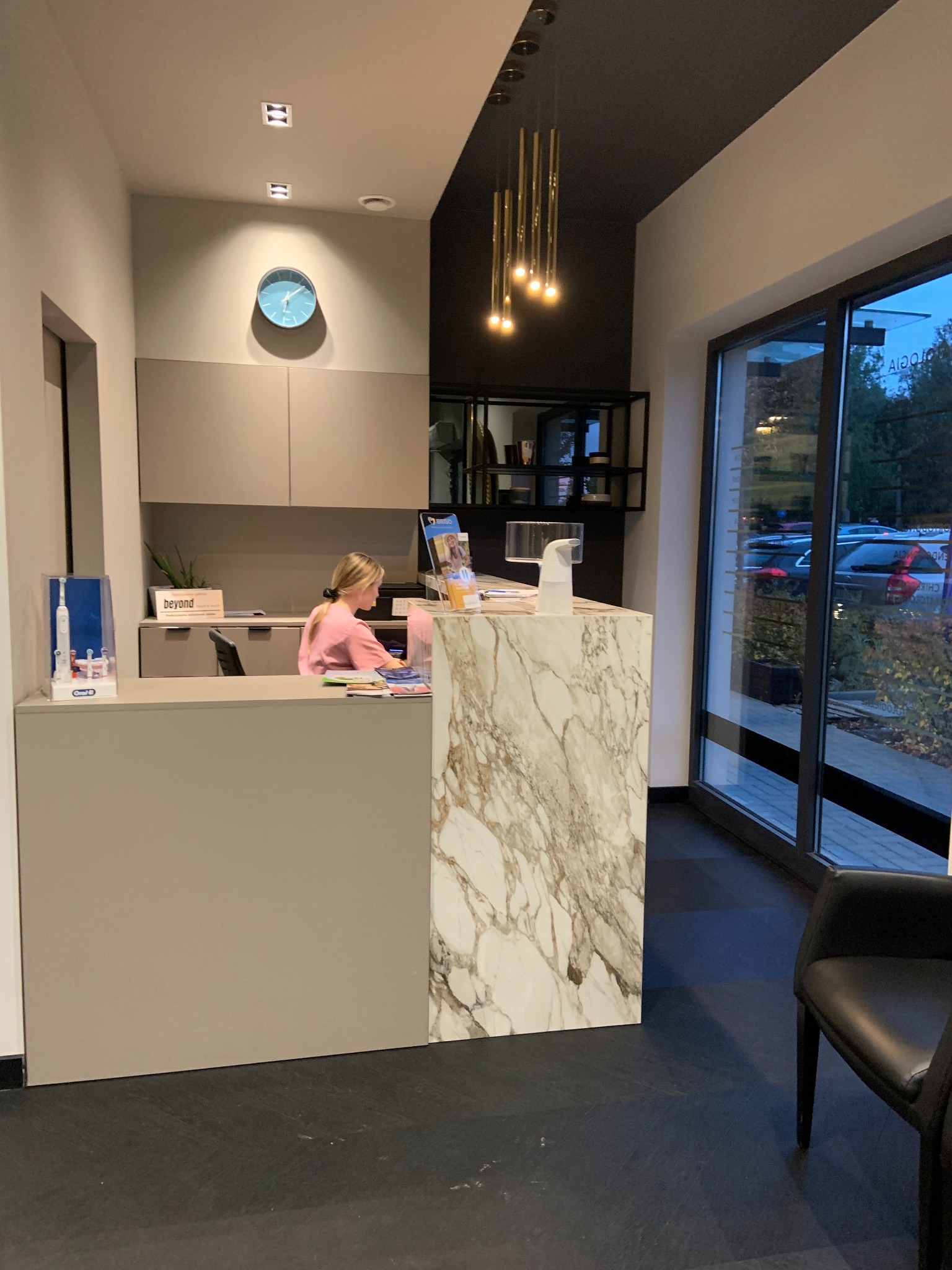 Recepcja w nowoczesnej klinice z marmurowym blatem, widoczna pracująca recepcjonistka, ekspozycja produktów do higieny jamy ustnej oraz widok na zewnątrz przez duże okno.