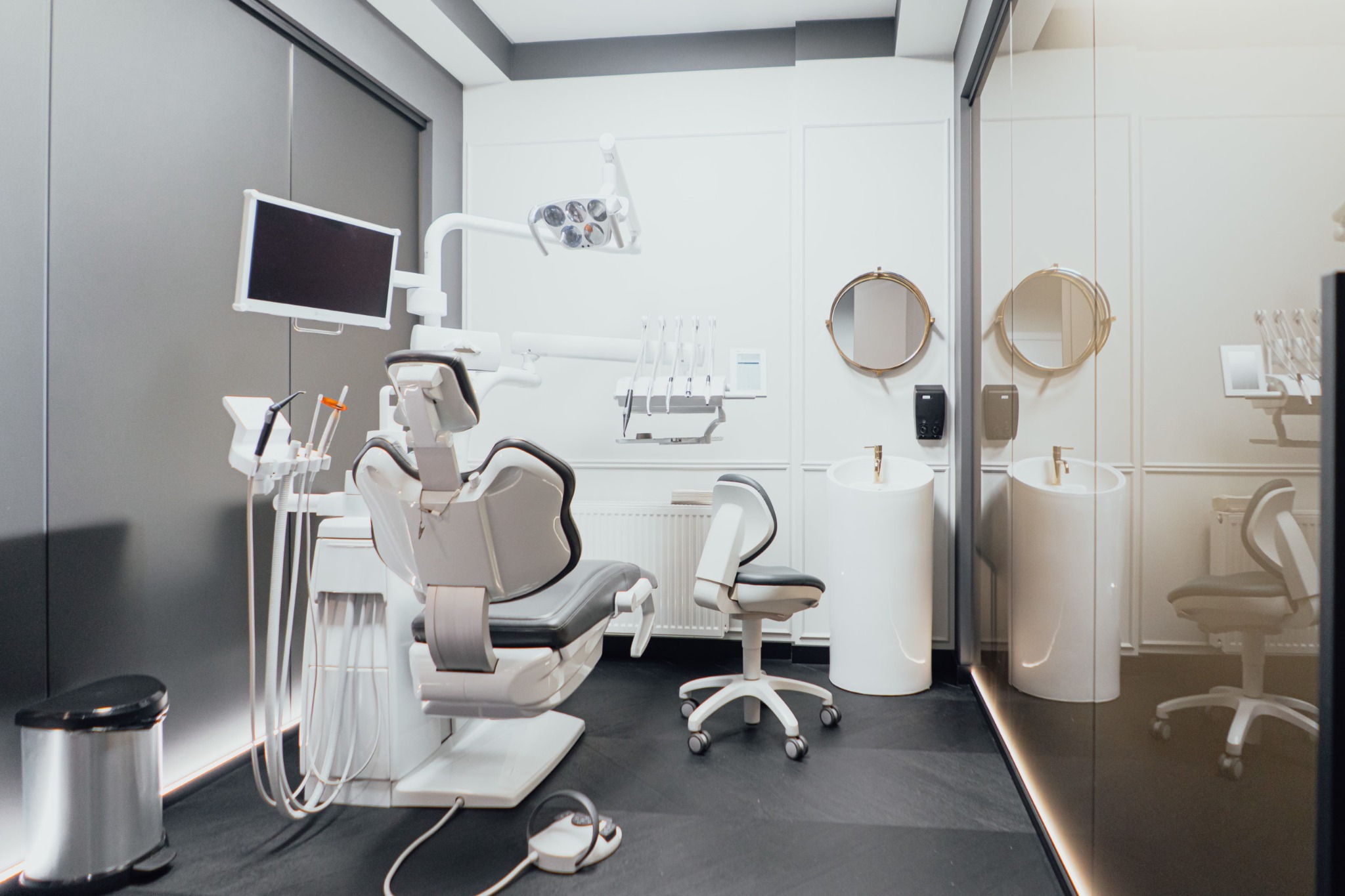 Nowoczesny gabinet stomatologiczny z szarymi ścianami, białymi meblami i sprzętem, widoczne krzesło dentystyczne, lampa zabiegowa, monitory i umywalki.