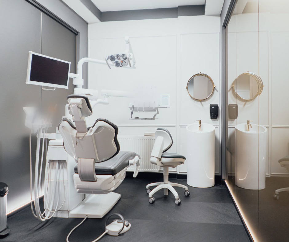 Nowoczesny gabinet dentystyczny z szarymi i białymi elementami, wyposażony w fotel dentystyczny, unit z monitorem, lampę zabiegową, umywalki na postumentach z okrągłymi lustrami oraz dodatkowe...