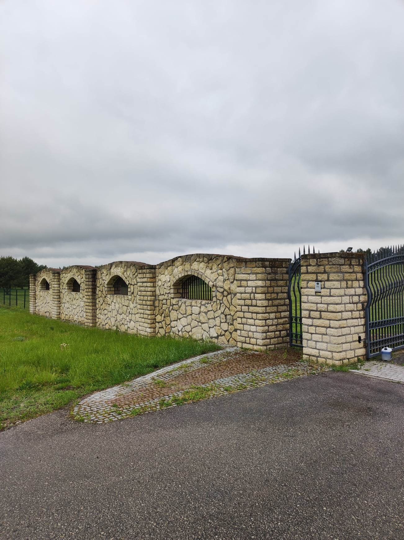 Kamienny mur z łukowymi oknami i metalową bramą wjazdową, widok z poziomu drogi asfaltowej w pochmurny dzień.