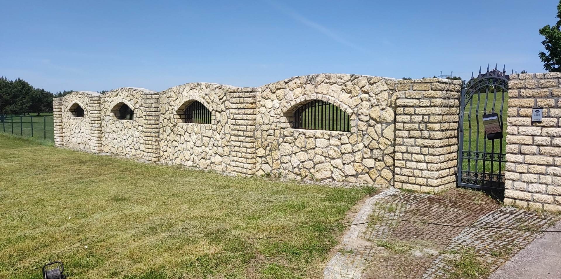 Kamienny mur z łukowymi oknami z metalowymi kratami i metalowa brama wjazdowa, otoczone trawnikiem i drzewami w tle.