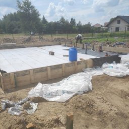 Magnum Best - Przygotowanie fundamentów pod budowę domu: drewniany szalunek, izolacja z białego styropianu, instalacja kanalizacyjna, widoczny pracownik w tle.