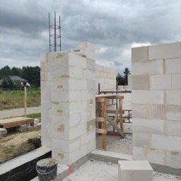 Magnum Best - Wznoszone ściany domu z białych bloczków betonowych, widoczne zbrojenie pionowe, drewniane rusztowanie w tle oraz wiadro na fundamencie, pochmurne niebo.