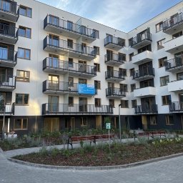 Magnum Best - Nowoczesny budynek wielorodzinny z białymi elewacjami, czarnymi balkonami i dużymi oknami, otoczony zagospodarowanym terenem zielonym z ławkami i chodnikiem z kostki brukowej.