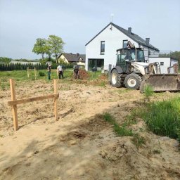 Magnum Best - Prace ziemne na działce budowlanej: traktor Terex wyrównuje teren pod budowę domu, w tle widoczny biały budynek w stylu nowoczesnym oraz grupa robotników przy wyznaczaniu granic.