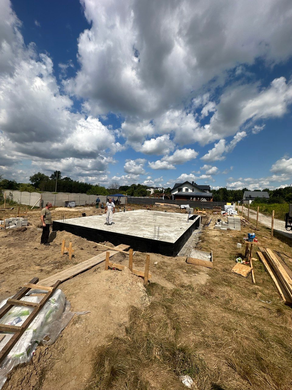 Fundament płyty betonowej z hydroizolacją, widoczni pracownicy budowlani, drewniane pomosty i materiały budowlane na działce z widokiem na dom w tle, błękitne niebo z białymi chmurami.