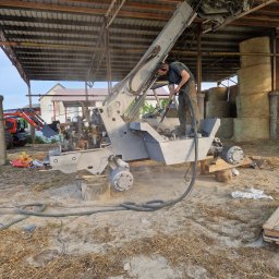 Fresz Natalia Loc - Pracownik w słuchawkach i okularach ochronnych piaskuje podwozie maszyny rolniczej na zewnątrz, przy stogu siana i pod zadaszeniem w słoneczny dzień.