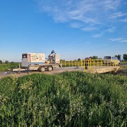 Fresz Natalia Loc - Mobilna piaskarka EKO na przyczepie, zaparkowana obok żółtej barierki mostu; pracownik w masce ochronnej przygotowuje się do piaskowania barierki, widoczna trawa i błękitne niebo.
