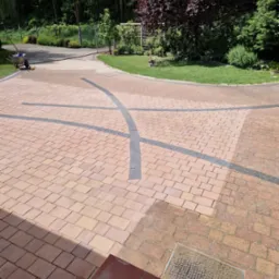 Czysta kostka brukowa w kolorze ceglastym z dekoracyjnymi wstawkami z szarej kostki, widok z góry, częściowo w cieniu schodów wejściowych.