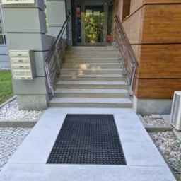 Zewnętrzne schody wejściowe do budynku biurowego z metalowymi poręczami i wycieraczką przed drzwiami.