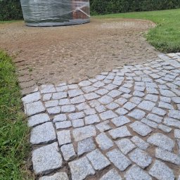 Fresz Natalia Loc - Fragment ścieżki z kostki brukowej w trakcie układania, z widocznym obszarem podsypki i zapakowaną w folię beczką w tle.