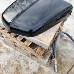 Fresz Natalia Loc - Błyszcząca, czarna pokrywa samochodu na drewnianej palecie, obok leży dysza do piaskowania. Widoczne odbicie drzew na powierzchni.