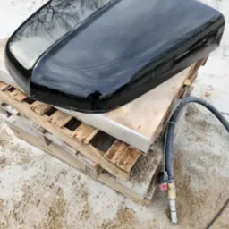 Błyszcząca, czarna pokrywa samochodu na drewnianej palecie, obok leży dysza do piaskowania. Widoczne odbicie drzew na powierzchni.