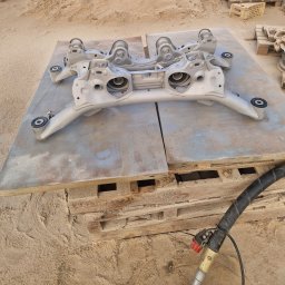 Fresz Natalia Loc - Szary, odnowiony element zawieszenia samochodowego leży na palecie, obok narzędzie do piaskowania. Tło to piasek. Detal po piaskowaniu i malowaniu.