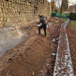 Fresz Natalia Loc - Czyszczenie kamiennej ściany strumieniem pod ciśnieniem przez pracownika w ogrodzie. Gabiony w tle.