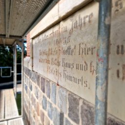 Fresz Natalia Loc - Fragment elewacji z kamienia i inskrypcją w gotyku, otoczony rusztowaniem. Renowacja zabytkowego muru z bliska, z widoczną fakturą kamienia i detalami napisu.