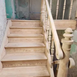 Drewniane schody z balustradą w surowym stanie, widoczne słoje drewna, fragment ściany z zieloną farbą w tle. Schody w trakcie renowacji lub montażu.