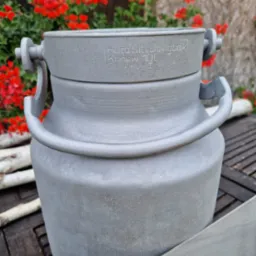 Szary, metalowy kanister z napisem 'Huta Silesia Rybnik Konew 10L' na drewnianym stole z gałązkami brzozy w tle oraz czerwonymi kwiatami.