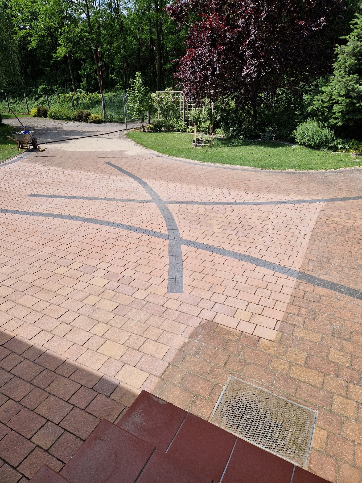 Czysta kostka brukowa w kolorze ceglastym z dekoracyjnymi wstawkami z szarej kostki, widok z góry, częściowo w cieniu schodów wejściowych.