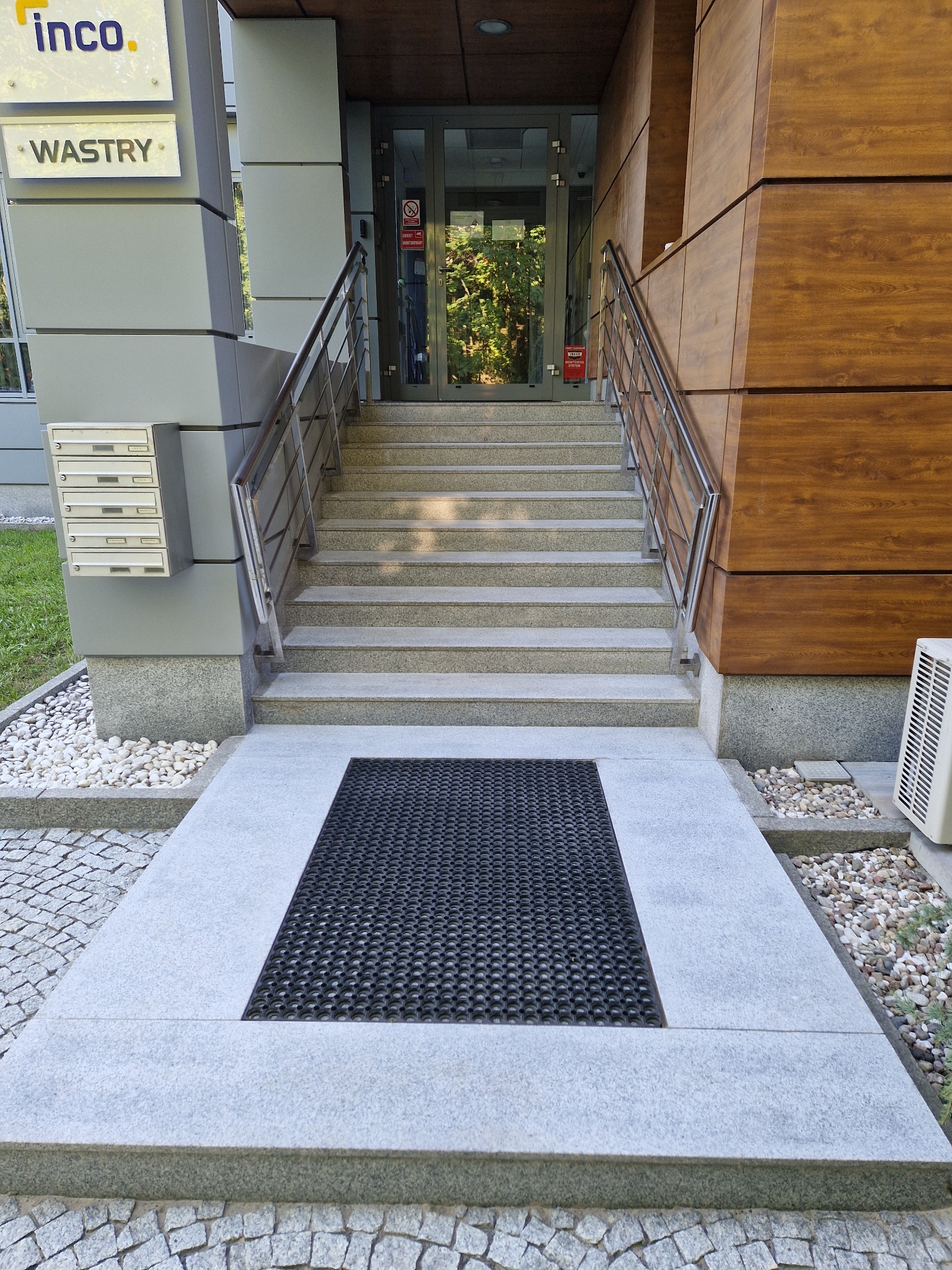 Zewnętrzne schody wejściowe do budynku biurowego z metalowymi poręczami i wycieraczką przed drzwiami.