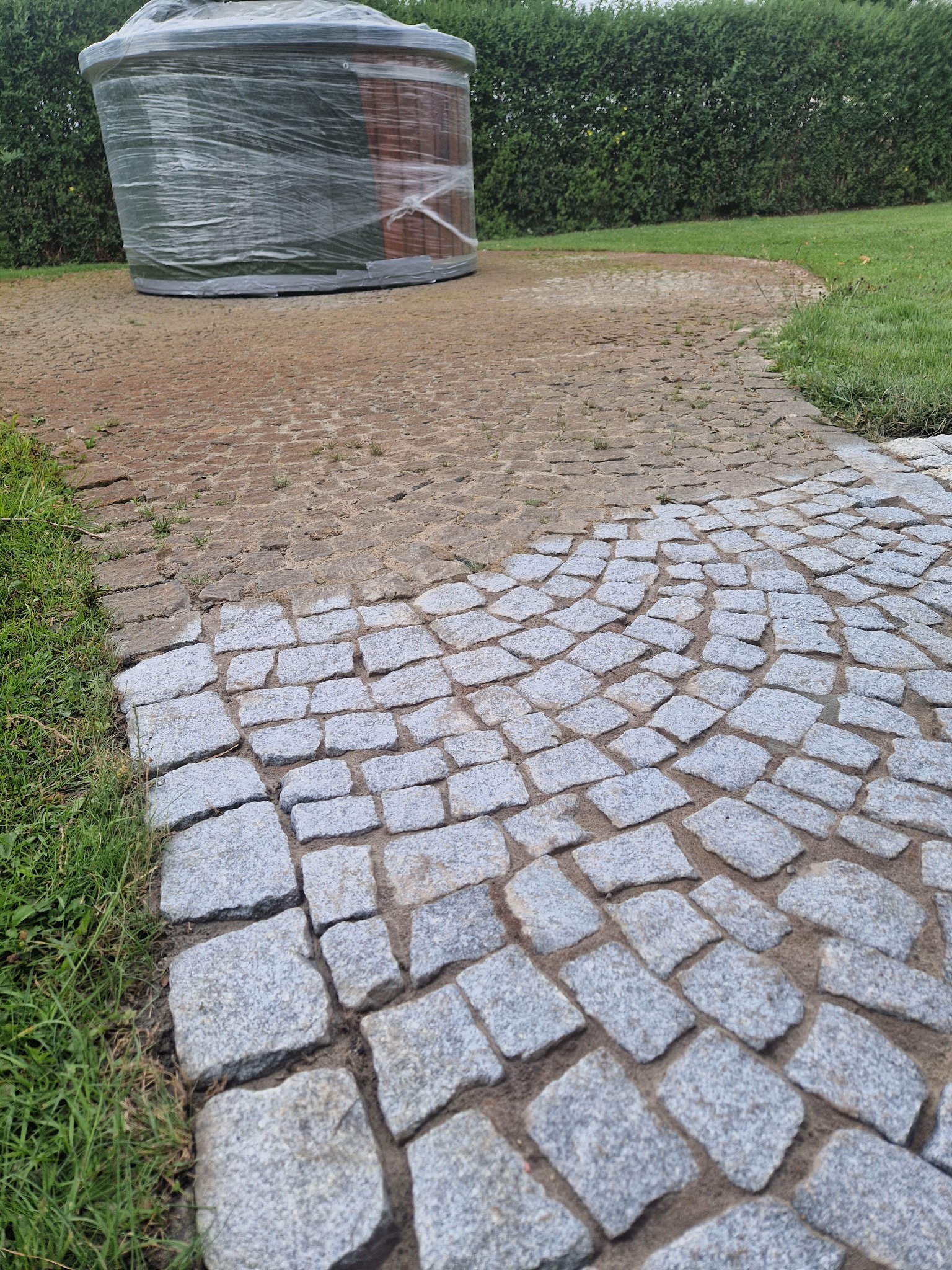 Fragment ścieżki z kostki brukowej w trakcie układania, z widocznym obszarem podsypki i zapakowaną w folię beczką w tle.