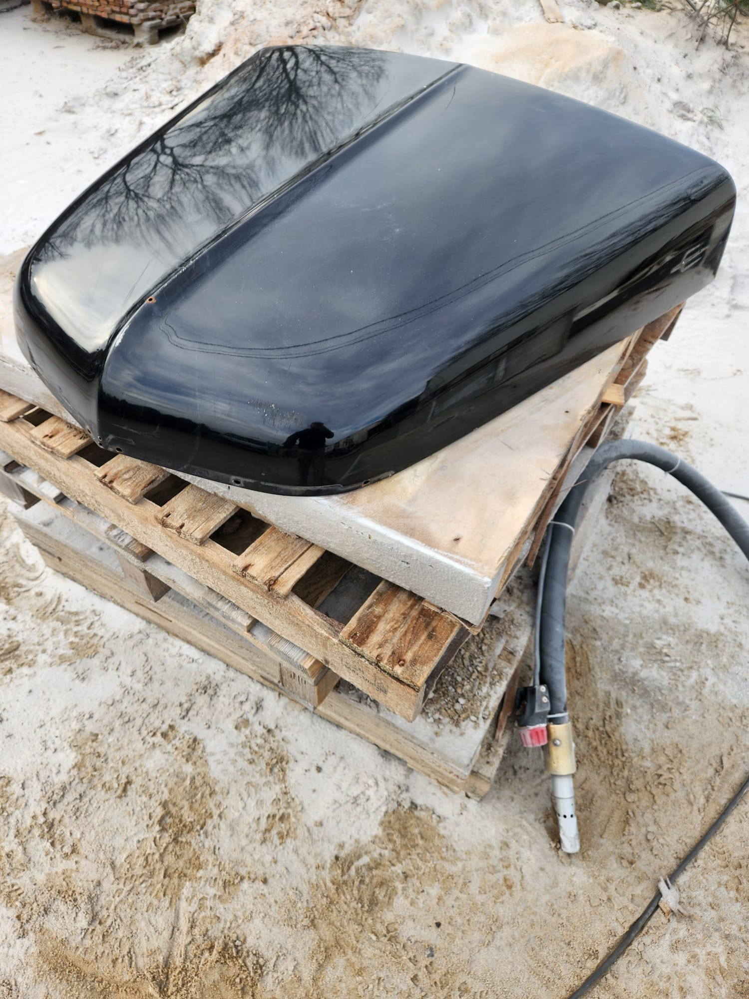 Błyszcząca, czarna pokrywa samochodu na drewnianej palecie, obok leży dysza do piaskowania. Widoczne odbicie drzew na powierzchni.