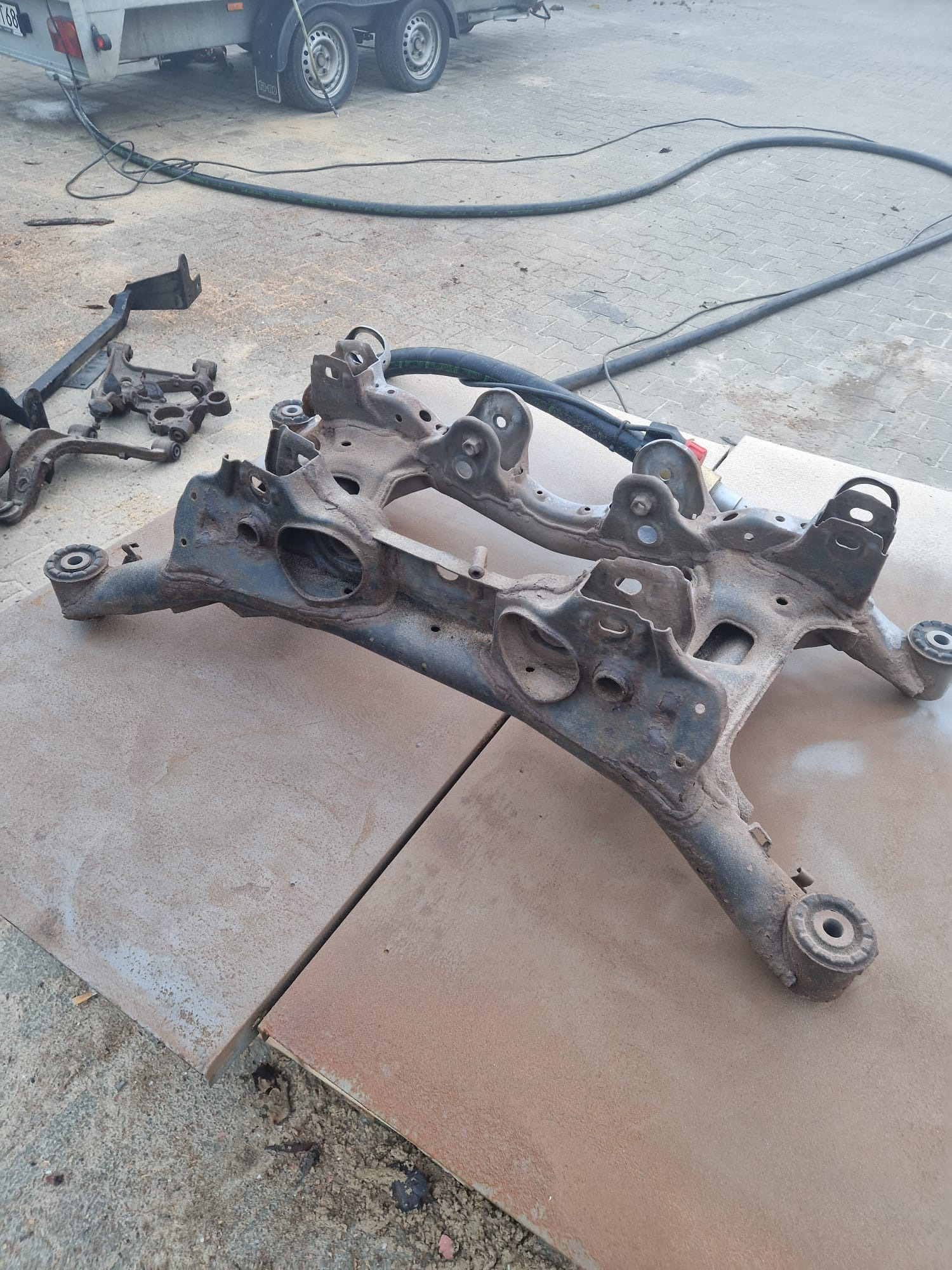 Dwie skorodowane ramy podwozia samochodowego leżące na płytach. W tle przyczepa i węże. Detal metalowych elementów przed renowacją.